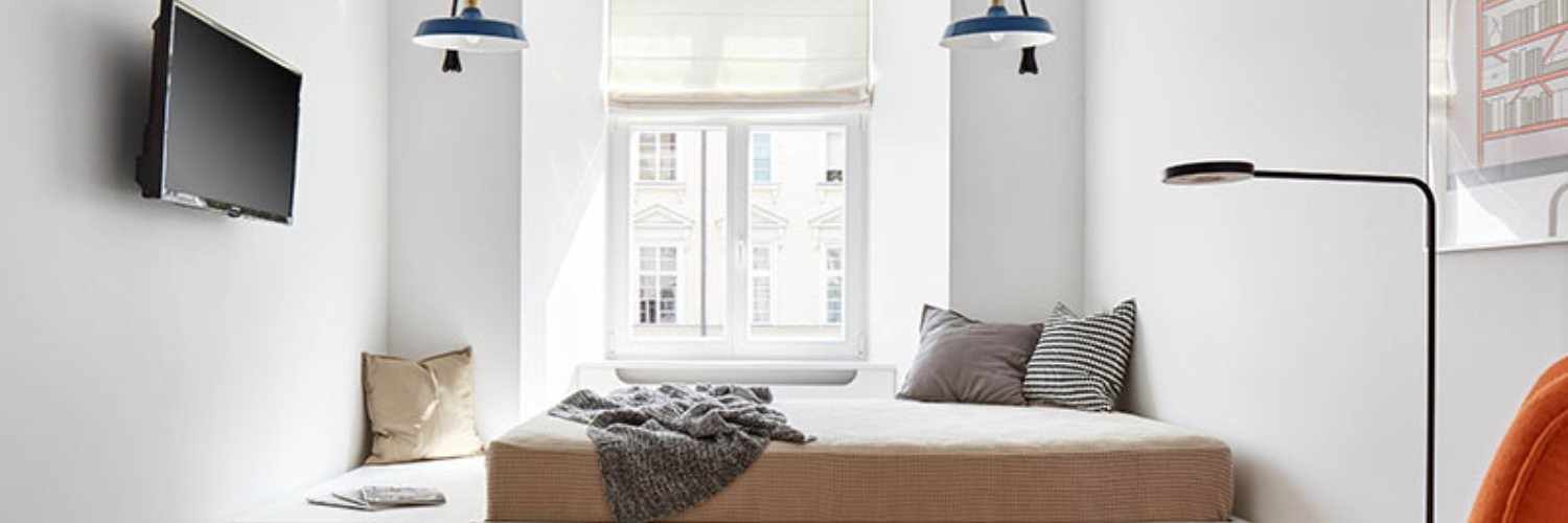 Minimalist bedroom with a cozy bed, layered pillows, neutral tones, a mounted TV, natural light, and stylish lighting fixtures.