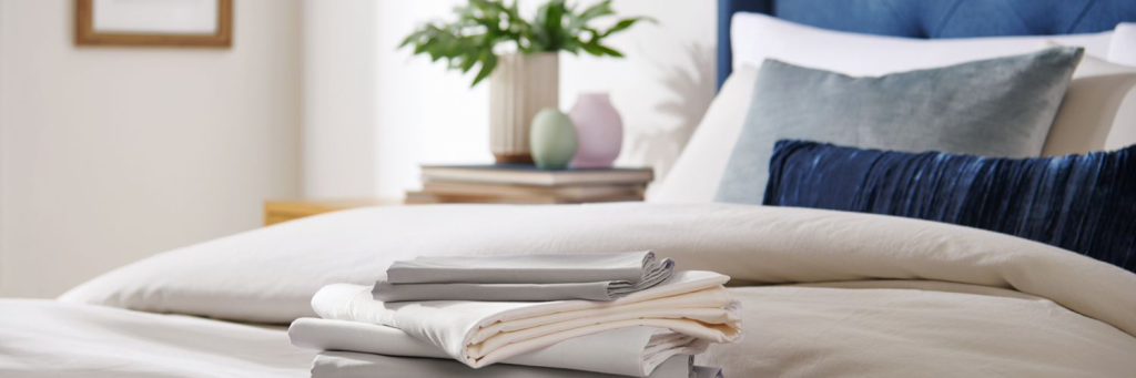 A neatly made bed featuring a stack of folded sheets in gray, cream, and white, with decorative pillows and a potted plant in the background.
