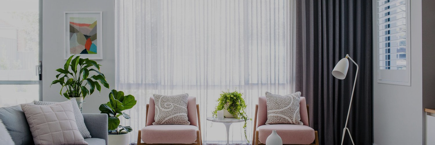 A modern living room with two pink chairs, a gray sofa, potted plants, and large curtains, creating a bright and inviting atmosphere.