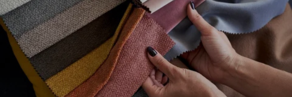 Two hands hold colorful upholstery fabric swatches, showcasing various textures and hues of textile materials arranged in a fan shape.