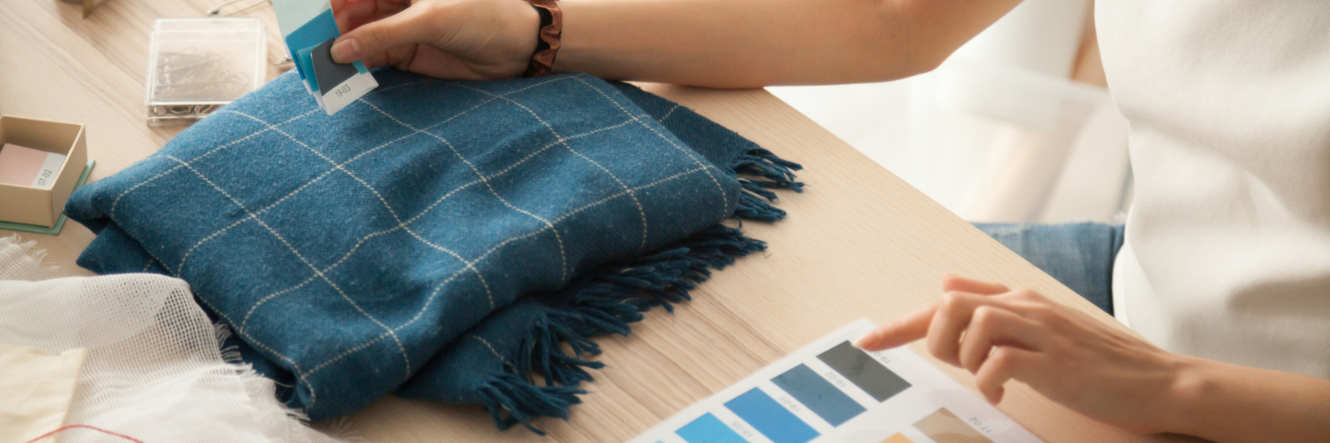 A person examines color swatches while comparing them to a blue plaid fabric laid on a wooden table.