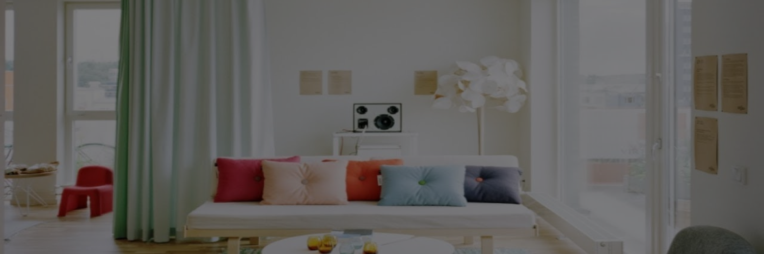 Bright living room with a white sofa adorned with colorful pillows, a minimalist speaker, and green curtains, bathed in natural light.