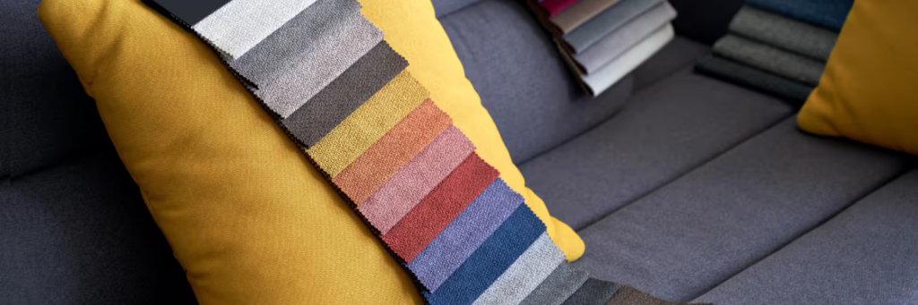 A vibrant color swatch display rests on a gray sofa with a bright yellow pillow, showcasing a variety of textile textures and hues.