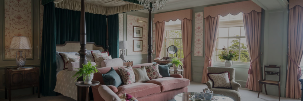Elegant bedroom featuring a four-poster bed, plush pink sofa, vintage decor, and large windows with drapes, showcasing a cozy ambiance.