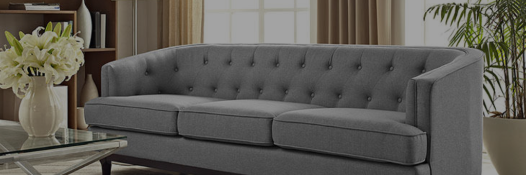 A stylish gray tufted sofa with three cushions, paired with a glass coffee table and a vase of white flowers in a bright living room.