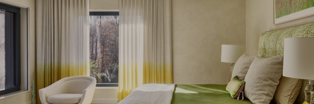 Modern bedroom featuring a green bedspread, yellow-dipped curtains, textured wall, and a cozy chair by the window overlooking nature.
