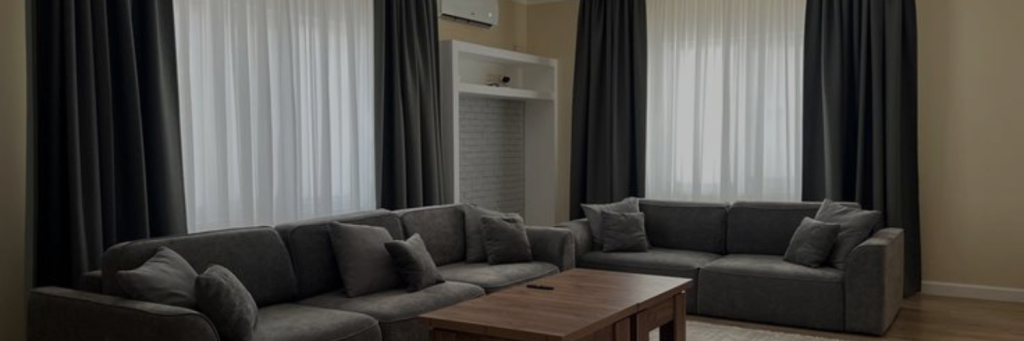 A cozy living room with two gray sectional sofas, a wooden coffee table, and large windows draped with dark curtains. The space feels modern and inviting.